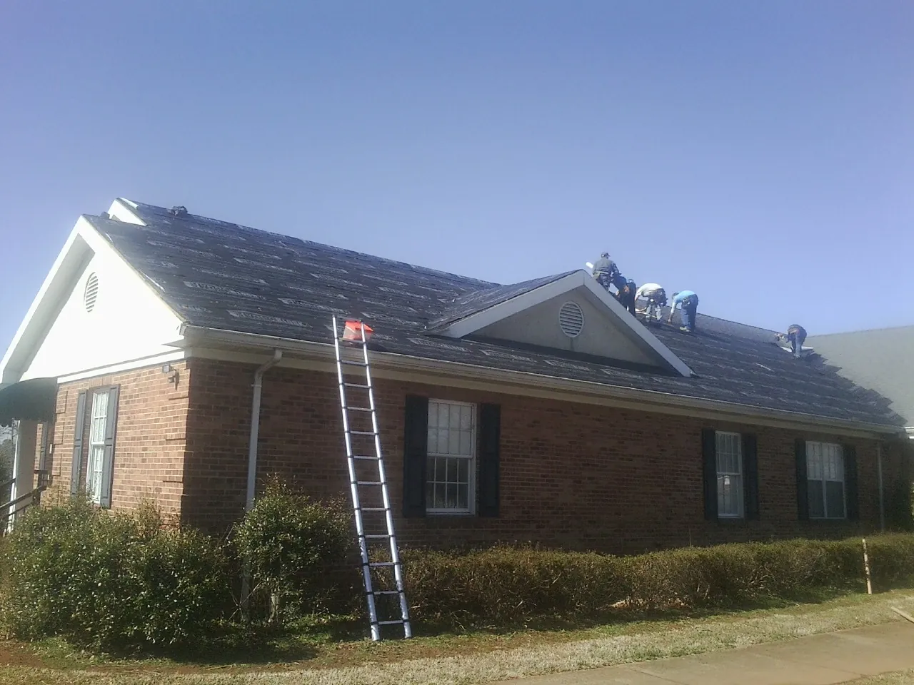Crew tearing shingles off a brick ranch roof with a ladder set — Metal Roof Repair work by our crew in Manitou Beach