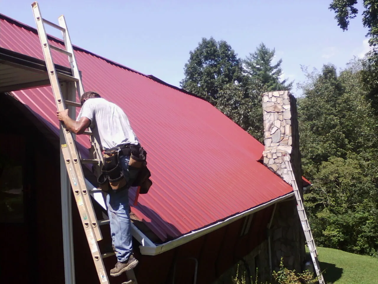 Roofer on a ladder beside a red metal roof and stone chimney — Soffit & Fascia Repair by Roofing Brothers in Manitou Beach